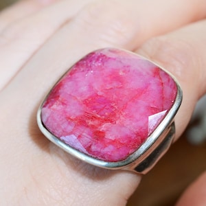 Könnte beinhalten: Ein Ring mit einem großen, quadratisch geschliffenen, facettierten, pinkfarbenen Edelstein, der in eine glatte, polierte silberfarbene Metallschiene gefasst ist. Der Stein zeigt eine natürliche, texturierte Oberfläche mit Magenta- und Rosatönen.