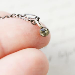 Könnte beinhalten: Nahaufnahme einer zarten Silberkette mit einem kleinen, runden, olivgrünen Edelsteinanhänger. Die Kette besteht aus kleinen, miteinander verbundenen Gliedern. Der Anhänger wird zwischen zwei Fingern gehalten, was die filigranen Details und die Handwerkskunst des Schmucks zeigt.