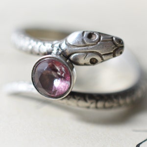 May include: A silver snake ring with a pink gemstone. The snake is coiled around the finger and the head is facing forward. The gemstone is set in the center of the snake's body.