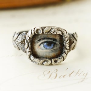 May include: A silver ring featuring a central square-shaped setting with rounded corners, containing a detailed miniature painting of a single blue eye. The metal band is decorated with a relief pattern of leaves along the sides and frame.