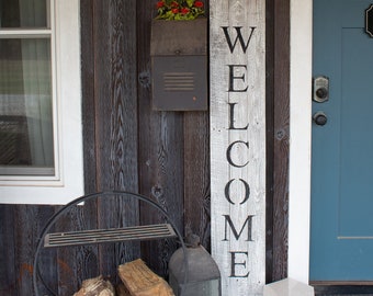 May include: A tall, rectangular, weathered gray wooden sign with the word WELCOME printed vertically in black capital letters. The sign is positioned upright against a dark brown vertical wood-paneled wall next to a blue door and a window.