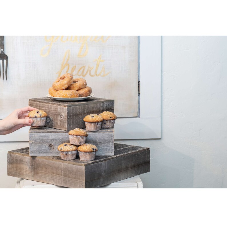 May include: A three-tiered wooden stand with a rustic finish, displaying a variety of baked goods. The top tier features a plate of glazed doughnuts, while the lower tiers hold blueberry muffins.