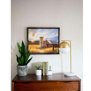May include: A framed canvas print of a red barn with a silo in a field of golden wheat. The sun is setting in the background, casting a warm glow over the scene. The print is hanging on a white wall above a wooden dresser with a lamp and plants.