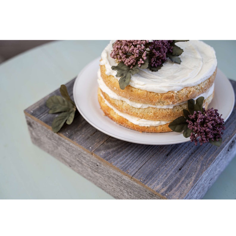 May include: A three-tiered cake with white frosting and pink flowers on top, sitting on a rustic wooden cake stand.