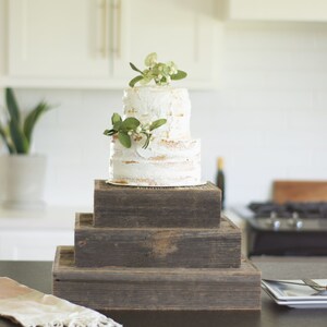 May include: A two-tiered white cake with buttercream frosting and green foliage on top, displayed on a rustic wooden cake stand with three tiers.