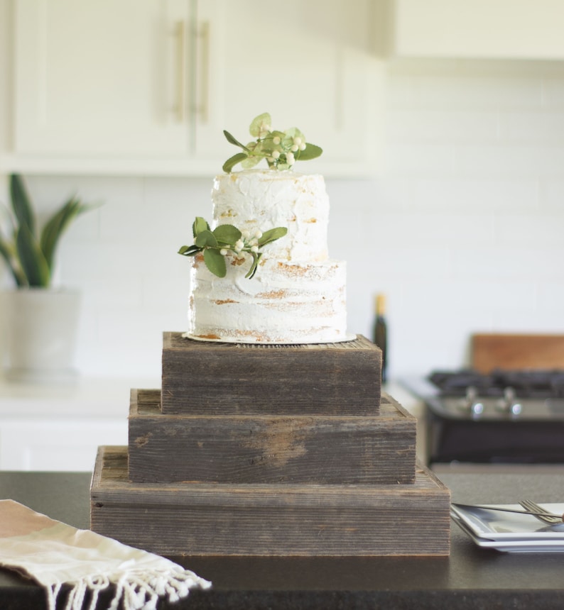 May include: A two-tiered white cake with buttercream frosting and green foliage on top, displayed on a rustic wooden cake stand with three tiers.