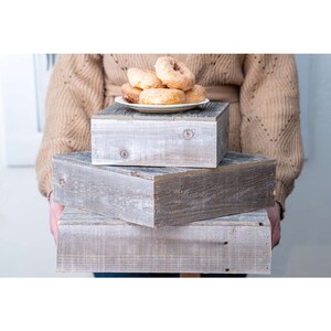 May include: A set of three wooden tiered display stands in a grey wash finish. The stands are stacked on top of each other and are being held by a person. The top stand has a white plate with a few glazed doughnuts on it.