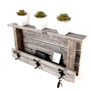 May include: A rustic wooden key holder with a shelf and three hooks. The shelf is made of reclaimed wood and has a gray finish. There are three small potted succulents on the shelf.