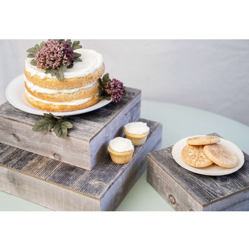 May include: Three rustic wooden cake stands with a white cake, cupcakes, and cookies on top. The stands are stacked on top of each other and are made of reclaimed wood.