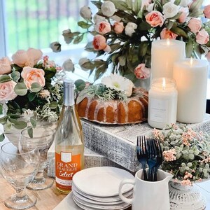 May include: A table setting with a white cake, white candles, a bottle of Grand Traverse Select Riesling wine, white plates, and white flowers.