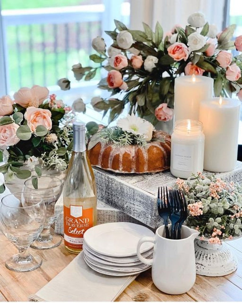 May include: A table setting with a white cake, white candles, a bottle of Grand Traverse Select Riesling wine, white plates, and white flowers.