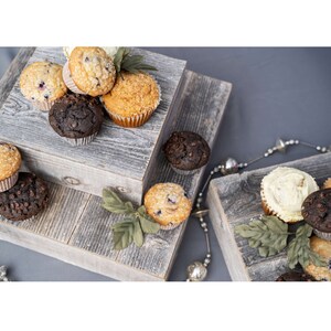 May include: A two-tiered wooden cake stand with a rustic grey finish. The stand is decorated with green leaves and features a variety of muffins, including chocolate chip, blueberry, and vanilla.