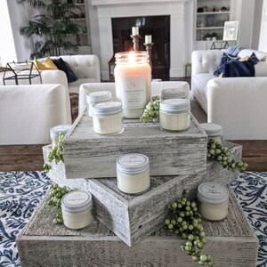 May include: A three-tiered wooden candle stand with a distressed white finish. The stand is decorated with greenery and features various sizes of glass candle jars with white lids. The largest jar has a label that reads "Peach Apricot".