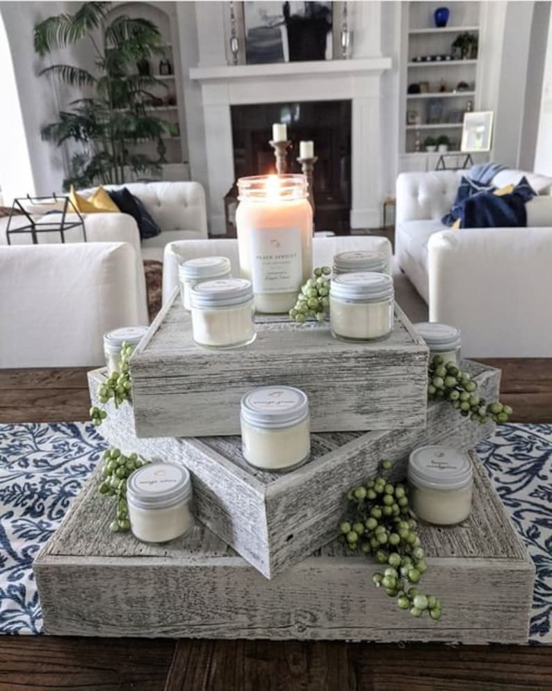 May include: A three-tiered wooden candle stand with a distressed white finish. The stand is decorated with greenery and features various sizes of glass candle jars with white lids. The largest jar has a label that reads "Peach Apricot".
