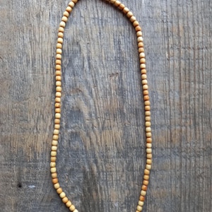 May include: A necklace made of small, round, light-colored wooden beads. The beads are strung together, forming a long strand. The necklace is displayed on a wooden surface with visible grain.