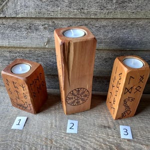 Op de afbeelding: Drie lichtbruine houten kandelaars van verschillende hoogtes, elk met een ronde opening aan de bovenkant voor een waxinelichtje. De houten oppervlakken zijn versierd met donkere, ingebrande runensymbolen, een kompasontwerp en een slangenmotief.
