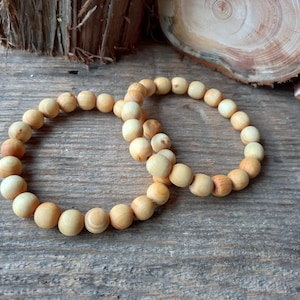 May include: Two wooden bead bracelets with a natural, light brown color. The beads are round and smooth, arranged in a circular pattern. The bracelets rest on a weathered wooden surface, with wood slices in the background.