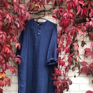Op de afbeelding: Een donkerblauwe linnen jurk met een knoopsluiting en drie knopen. De jurk heeft lange mouwen en een zijsplit aan elke kant. De jurk hangt aan een hanger tegen een witte bakstenen muur met rode klimop.