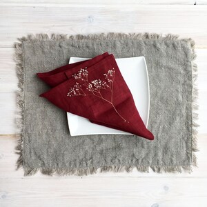 May include: A burgundy linen napkin folded into a triangle with a dried flower sprig on top. The napkin is placed on a gray linen placemat.