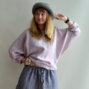 May include: A woman wearing a light pink linen top and a gray linen skirt. She is wearing a gray hat and has her hand on the brim of the hat.