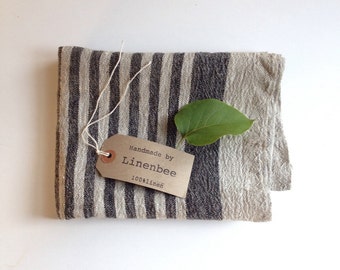 May include: A striped linen tea towel with a brown tag that says "Handmade by Linenbee 100%linen". A green leaf is on top of the towel.