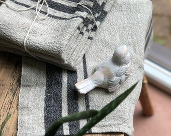 May include: A set of four gray linen tea towels with black stripes. The towels are folded and tied together with twine. A small white ceramic bird figurine is on top of the towels.
