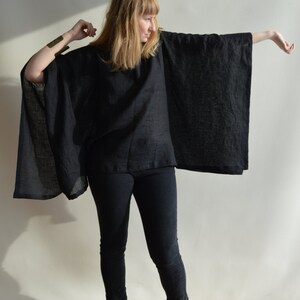 May include: A woman wearing a black linen poncho with long sleeves and a black pair of jeans. She is wearing brown leather shoes with a black trim.