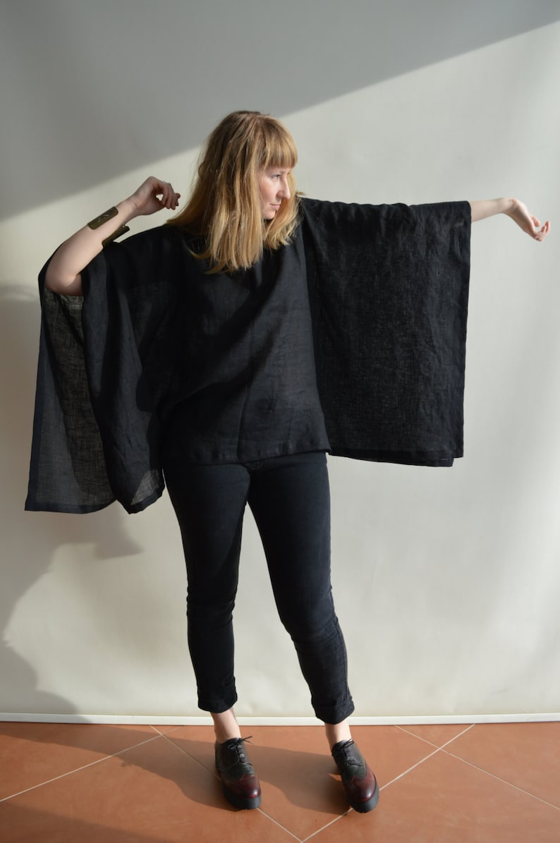 May include: A woman wearing a black linen poncho with long sleeves and a black pair of jeans. She is wearing brown leather shoes with a black trim.
