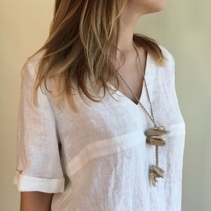 May include: A white linen shirt with a v-neck and short sleeves. The shirt features a simple design and is paired with a necklace made of driftwood and twine.