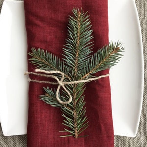 May include: A red linen napkin with a sprig of evergreen tied with twine. The napkin is folded and placed on a white plate.