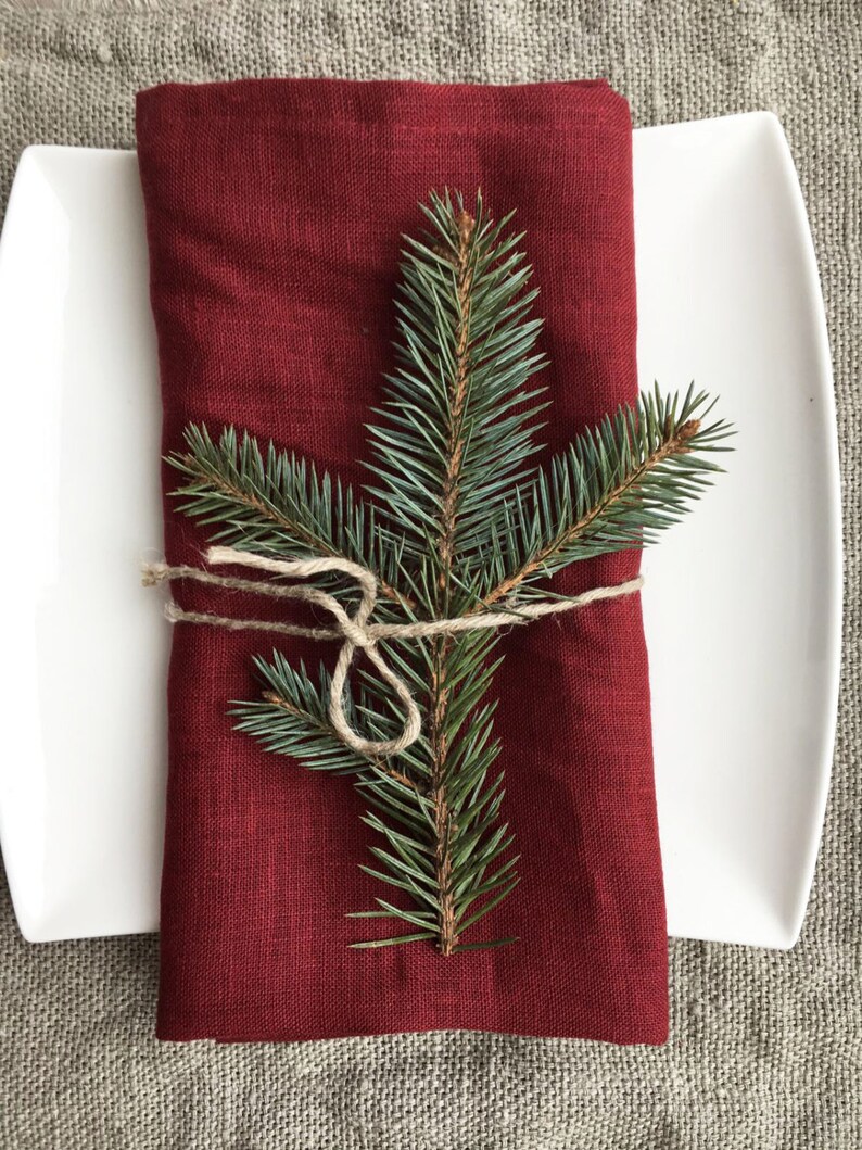 May include: A red linen napkin with a sprig of evergreen tied with twine. The napkin is folded and placed on a white plate.