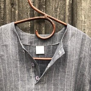 Op de afbeelding: Een grijs gestreept linnen shirt met een v-hals en knoopsluiting. Het shirt hangt aan een houten hanger.