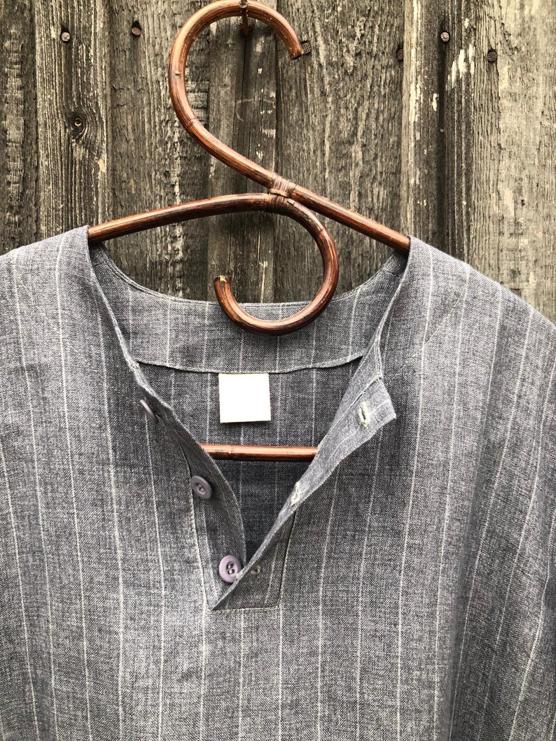 Op de afbeelding: Een grijs gestreept linnen shirt met een v-hals en knoopsluiting. Het shirt hangt aan een houten hanger.