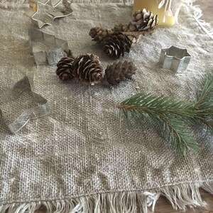 May include: A rustic linen table runner with a frayed edge, featuring a collection of metal cookie cutters in the shapes of a star, a tree, and a flower. Pine cones and sprigs of evergreen are scattered across the runner, creating a festive and natural scene.