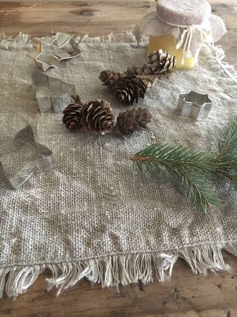 May include: A rustic linen table runner with a frayed edge, featuring a collection of metal cookie cutters in the shapes of a star, a tree, and a flower. Pine cones and sprigs of evergreen are scattered across the runner, creating a festive and natural scene.