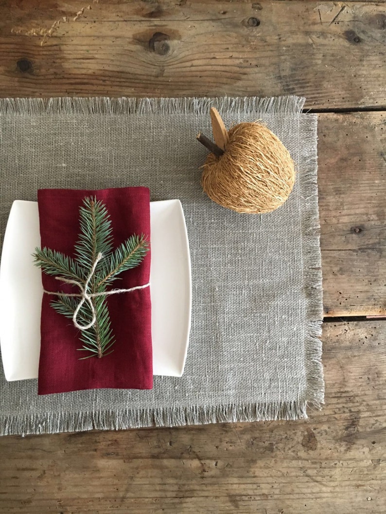 May include: A red linen napkin with a sprig of pine tied with twine, placed on a white plate and a gray linen placemat with frayed edges. A decorative woven apple sits on the placemat.