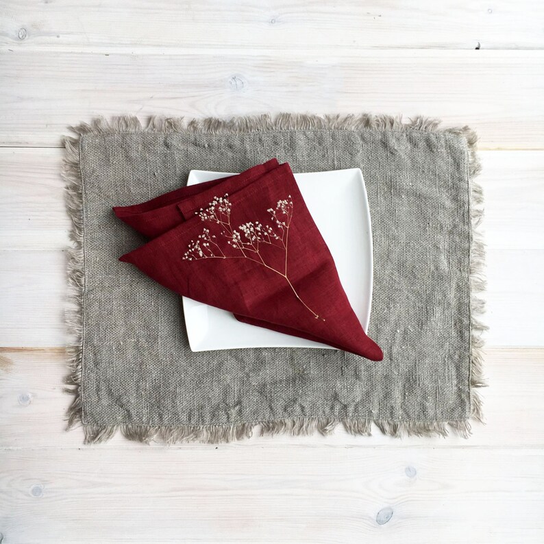 May include: A burgundy linen napkin folded into a triangle with a dried flower sprig on top. The napkin is placed on a gray linen placemat.