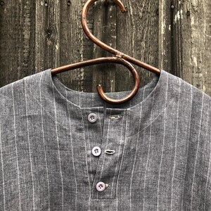 Op de afbeelding: Een grijs gestreept linnen shirt met een knoopsluiting. Het shirt hangt aan een houten hanger.