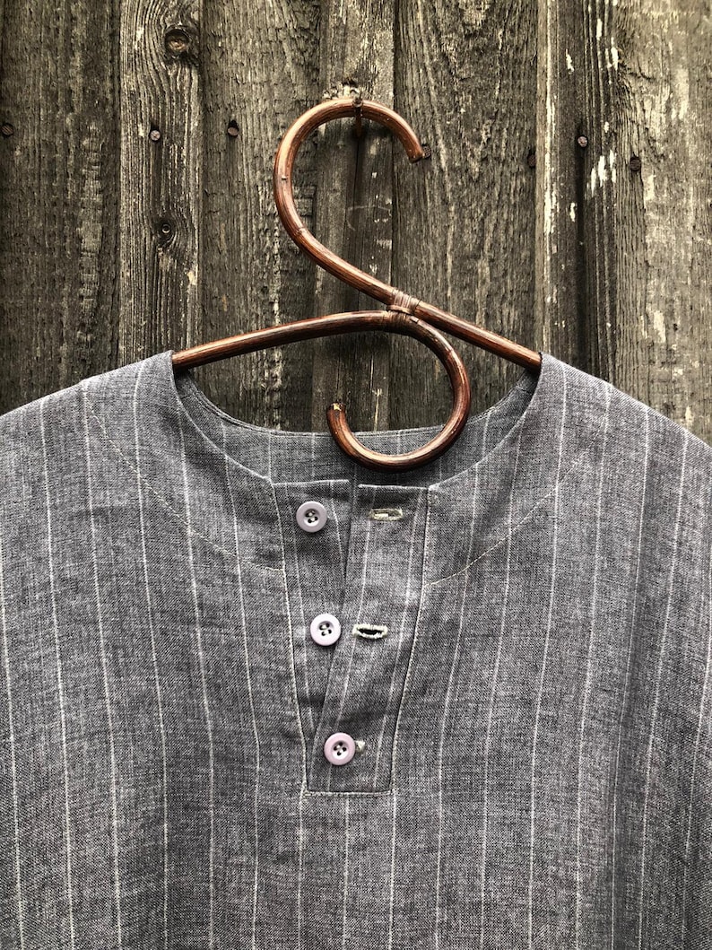 Op de afbeelding: Een grijs gestreept linnen shirt met een knoopsluiting. Het shirt hangt aan een houten hanger.