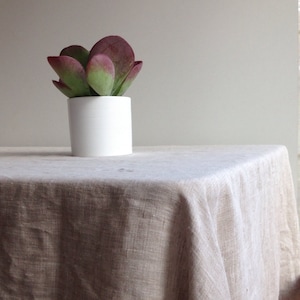 Op de afbeelding: Een witte keramische pot met een geribbelde textuur bevat een vetplant met groene en paarse bladeren. De pot staat op een lichtbruin linnen tafelkleed.