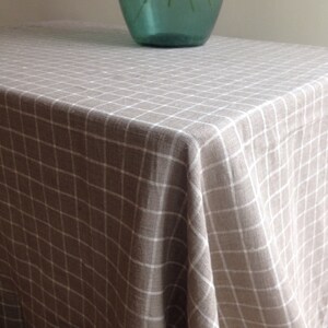 May include: A beige linen tablecloth with a white checkered pattern. The tablecloth is draped over a table and has a slight wrinkle in the center.