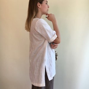 May include: A white linen tunic top with a side slit and short sleeves. The tunic is worn with brown pants.
