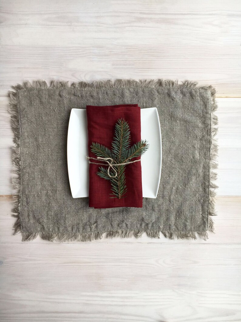 May include: A white plate with a red linen napkin tied with twine and a sprig of green pine needles on top. The plate is on a gray linen placemat.