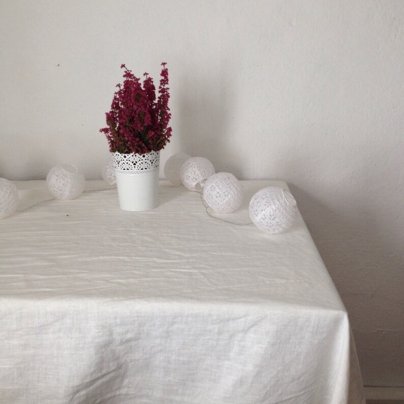 May include: A white tablecloth with a white vase filled with pink heather flowers and a string of white cotton ball lights.
