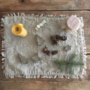 May include: A rustic linen placemat with a yellow candle, pine cones, and metal cookie cutters shaped like stars and Christmas trees.