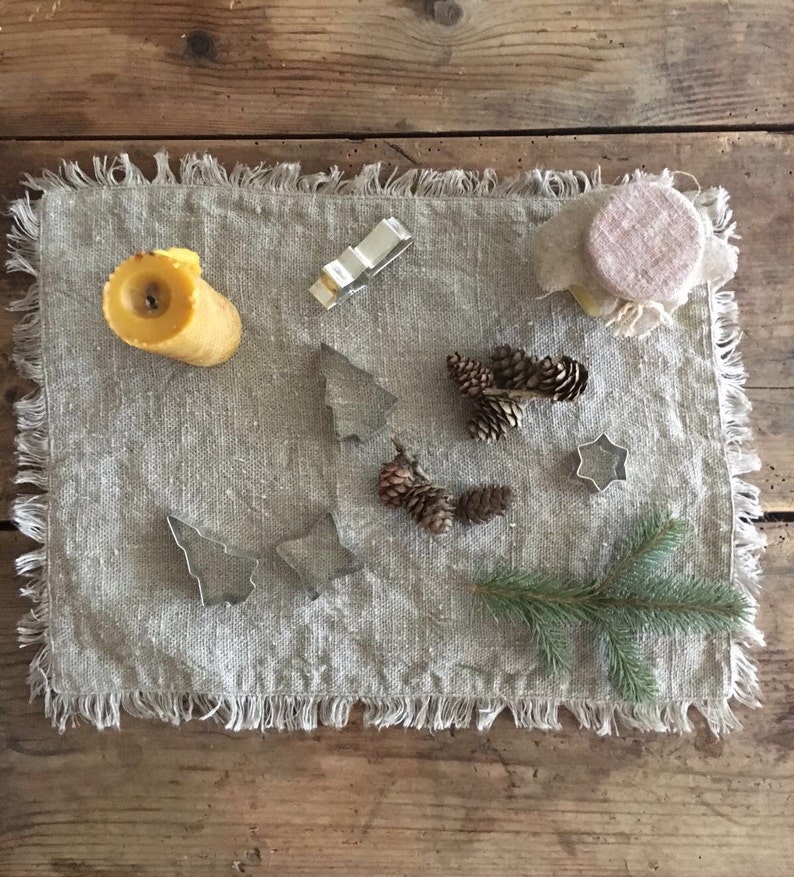 May include: A rustic linen placemat with a yellow candle, pine cones, and metal cookie cutters shaped like stars and Christmas trees.