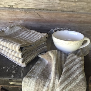 May include: A stack of brown and white striped linen tea towels with a white ceramic cup with a decorative design on a wooden surface.