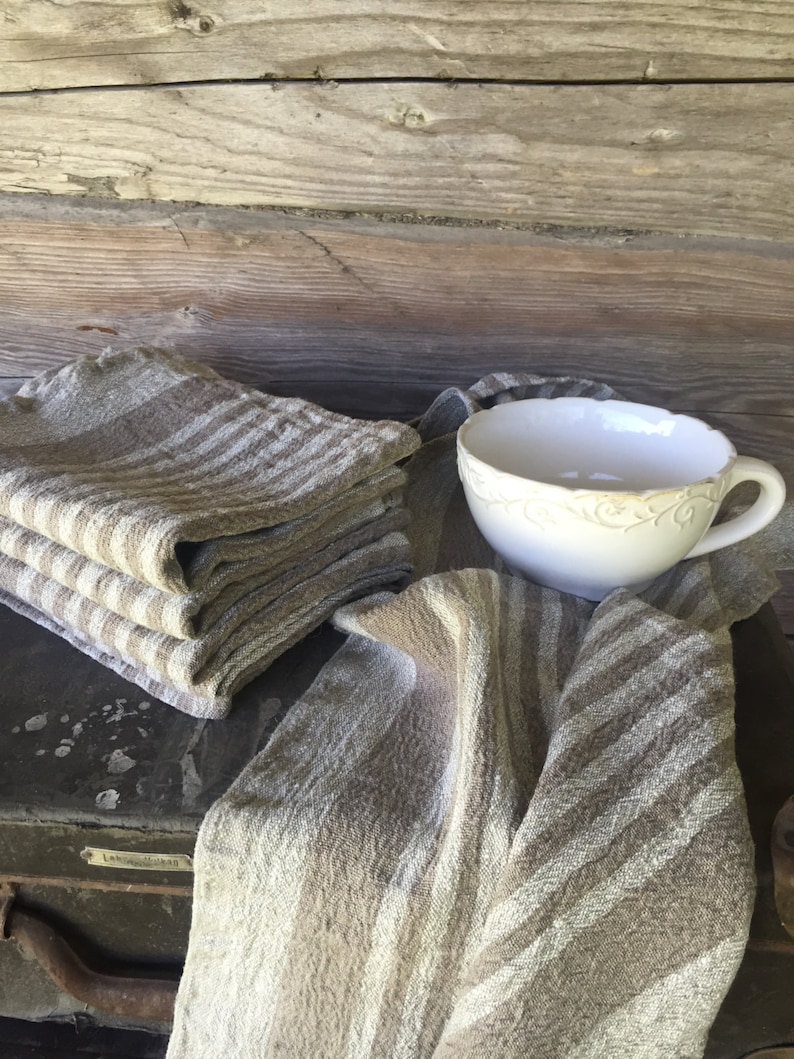 May include: A stack of brown and white striped linen tea towels with a white ceramic cup with a decorative design on a wooden surface.