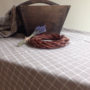 May include: A brown wooden box with a handle sits on a beige and white checkered tablecloth. A small bouquet of lavender flowers and a brown wicker wreath are in front of the box.