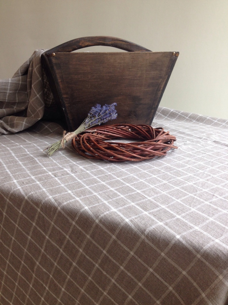 May include: A brown wooden box with a handle sits on a beige and white checkered tablecloth. A small bouquet of lavender flowers and a brown wicker wreath are in front of the box.